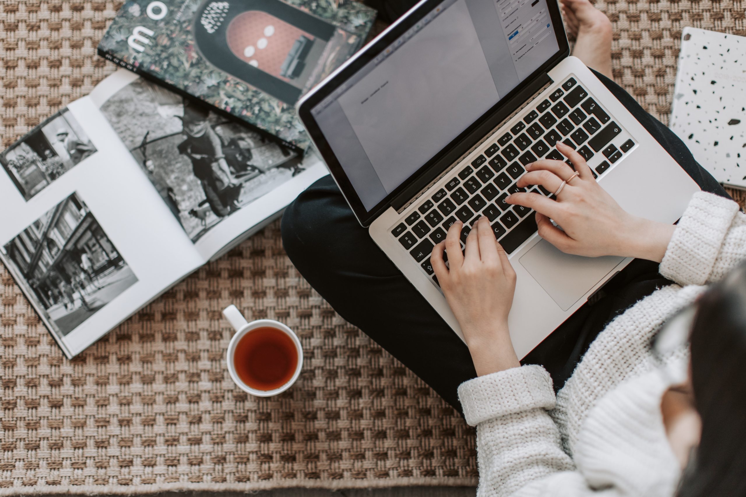 Woman typing on a laptop with a cup of tea and magazine articles next to her