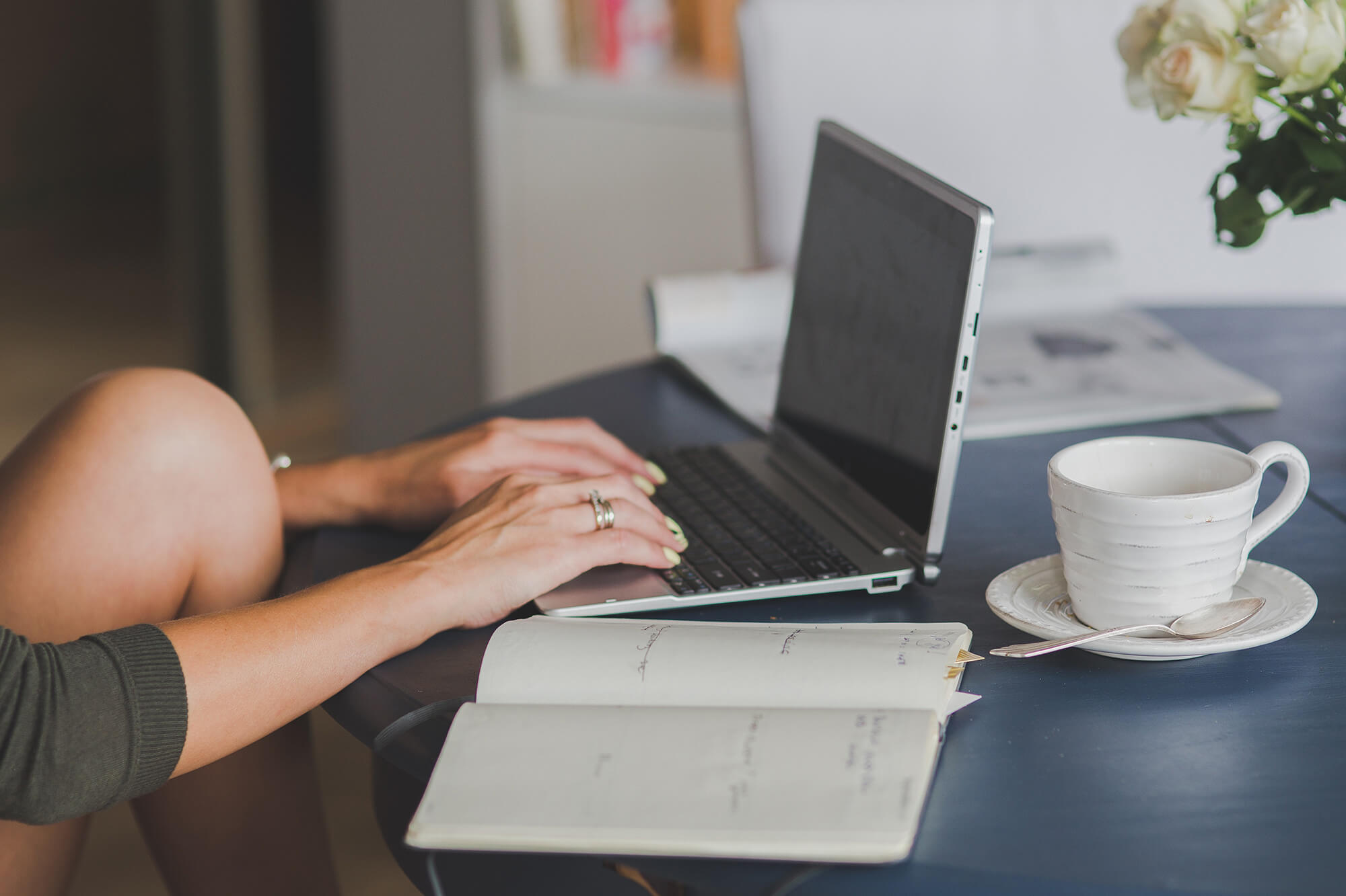 a woman types on a laptop while an open notebook and a coffee mug is next to her