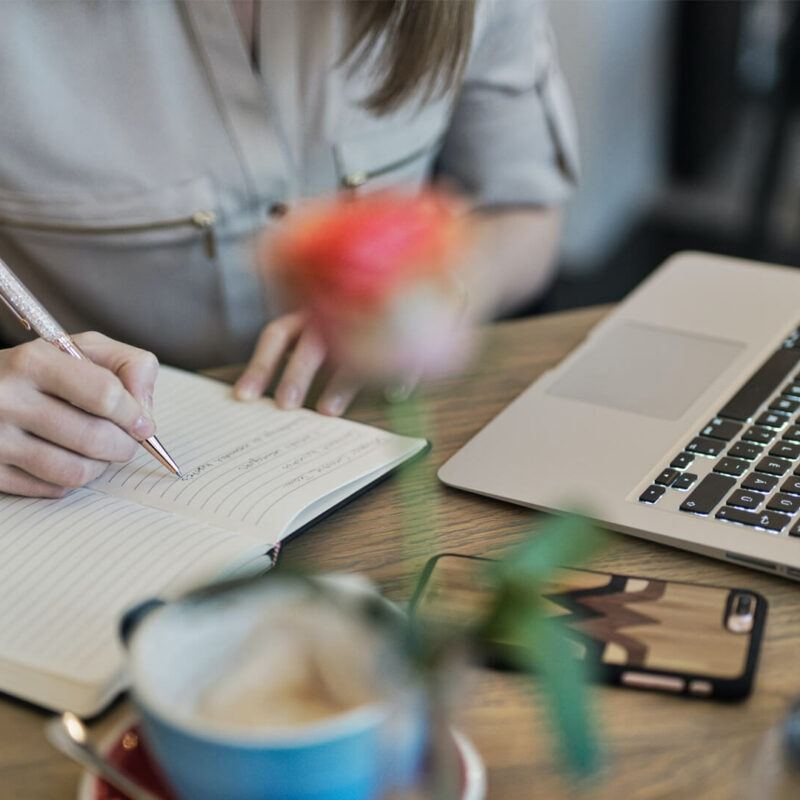 A person writes in a notebook next to their laptop on a wooden desk