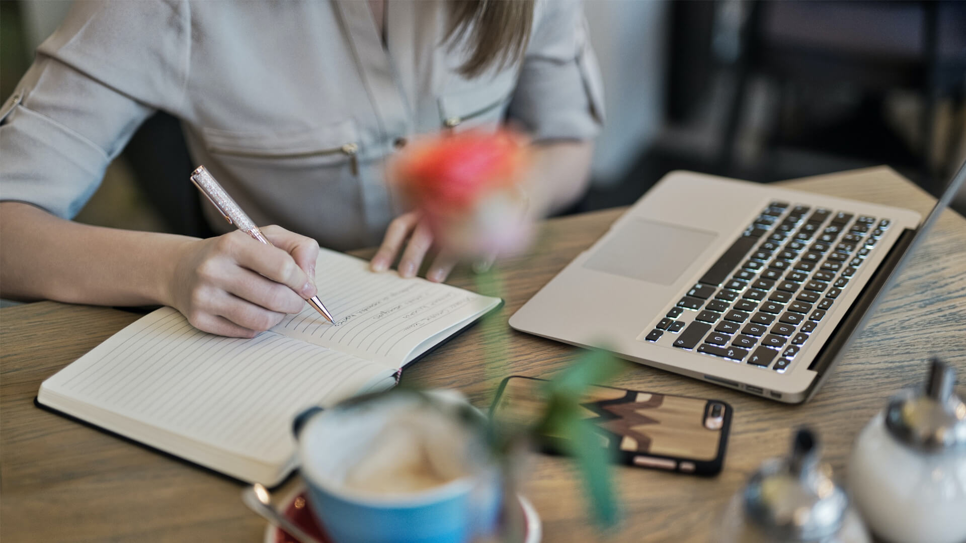 A person writes in a notebook next to their laptop on a wooden desk