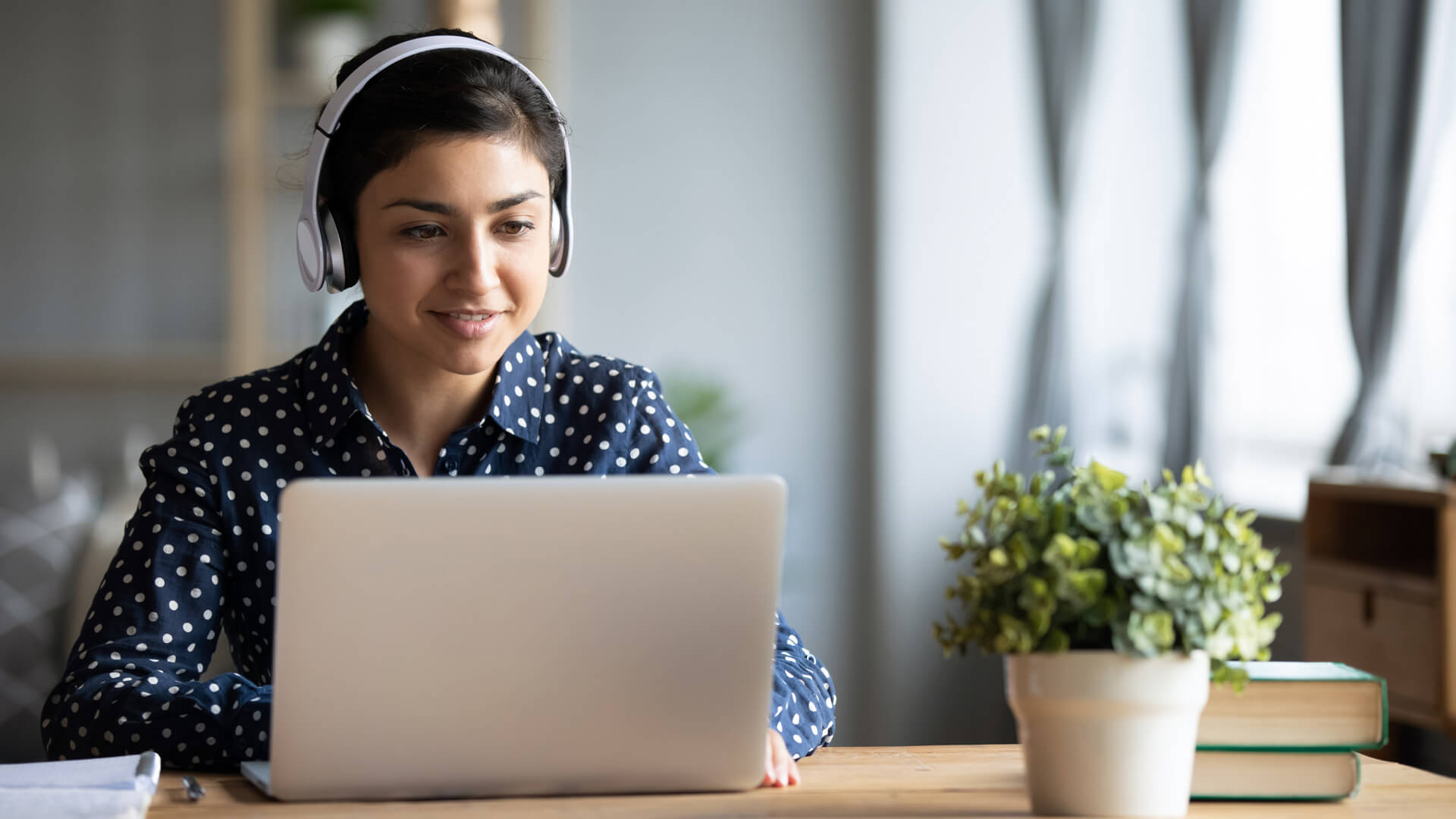 A woman wearing headphones sits in front of a laptop on a wooden desk. There is a small white pot with a plant and two stacked books on the desk. The woman is wearing a blue button up shirt with white dots.