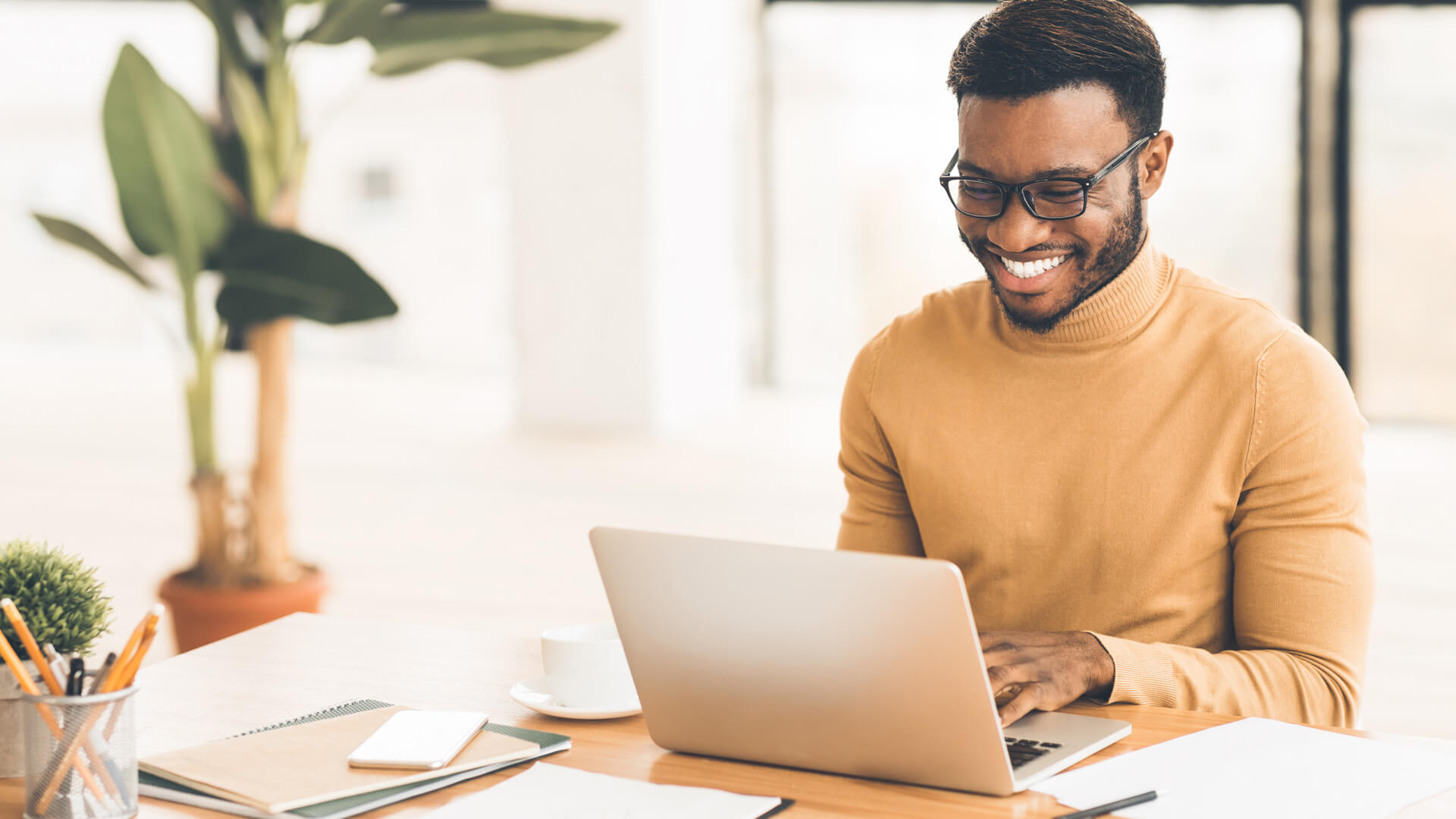 A Black man smiles as he types on a laptop on a wooden desk. A plant grows in the background. he wears glasses and a beige turtleneck.