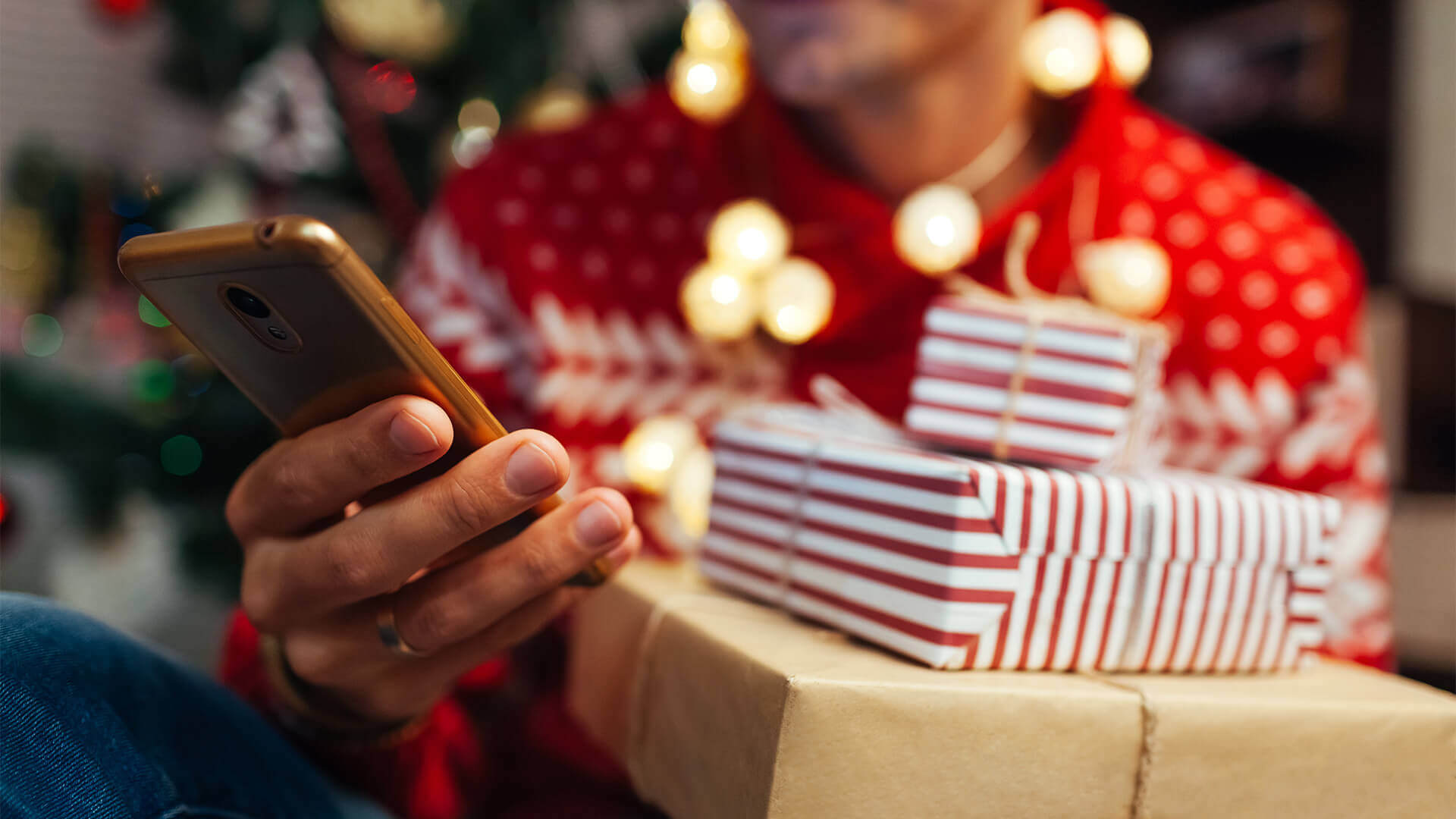 A person wearing a red and white holiday sweater holds three gifts in one hand and a phone in the other; the top two gifts are red and white striped, while the bottom gift is a plain beige wrapping paper. The person is sitting in front of a Christmas tree.