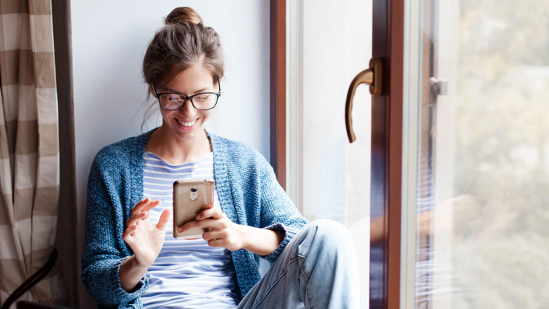 A woman smiles as she sits by a window and looks at her phone