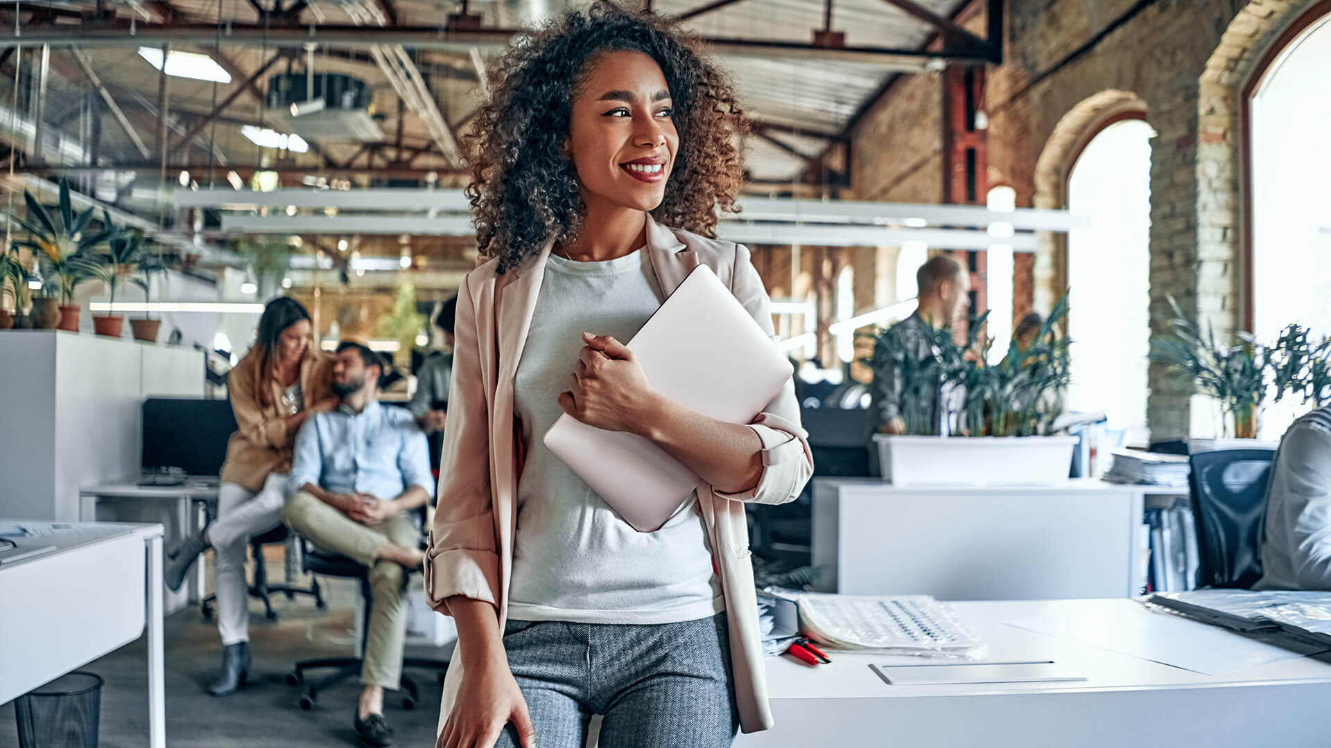 A woman with curly hair holds a laptop and looks off to the side in an office