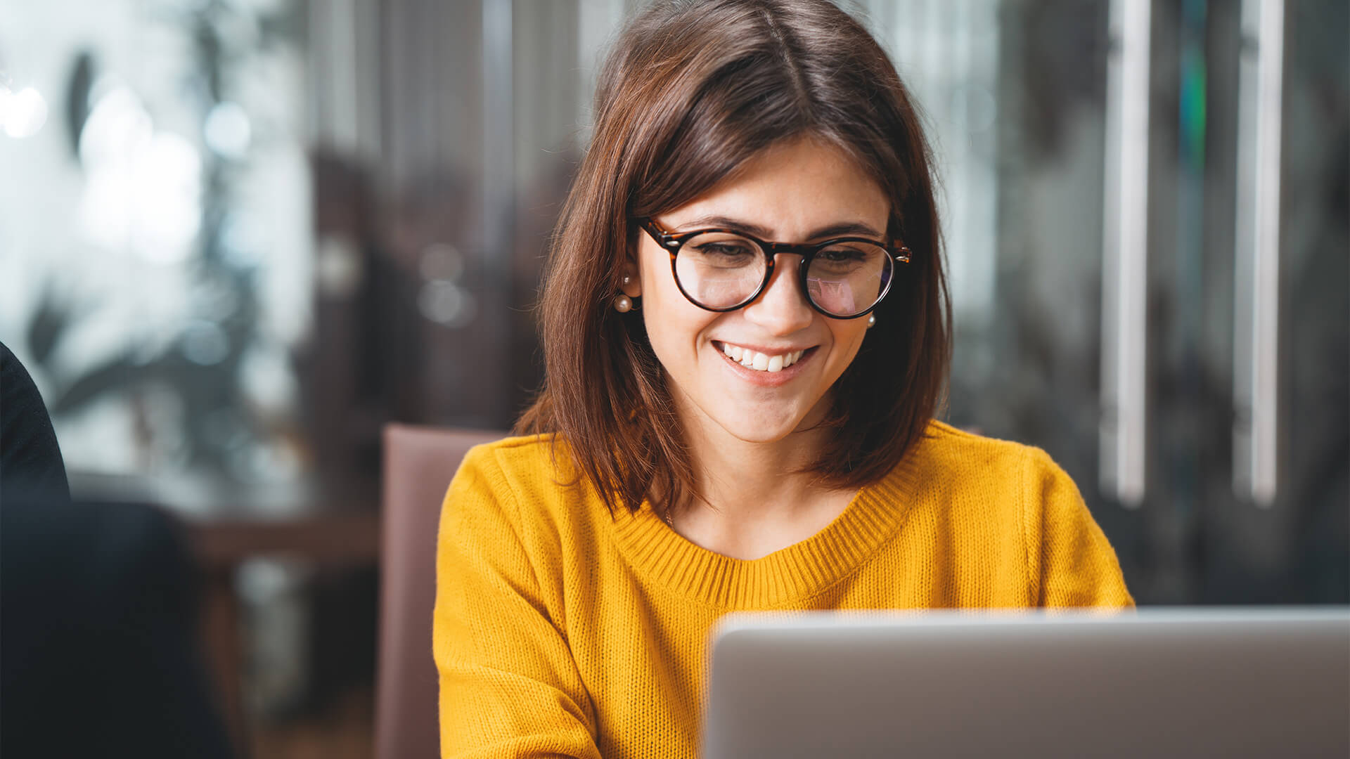 A woman with brown hair and glasses and a yellow sweater types on a laptop while smiling