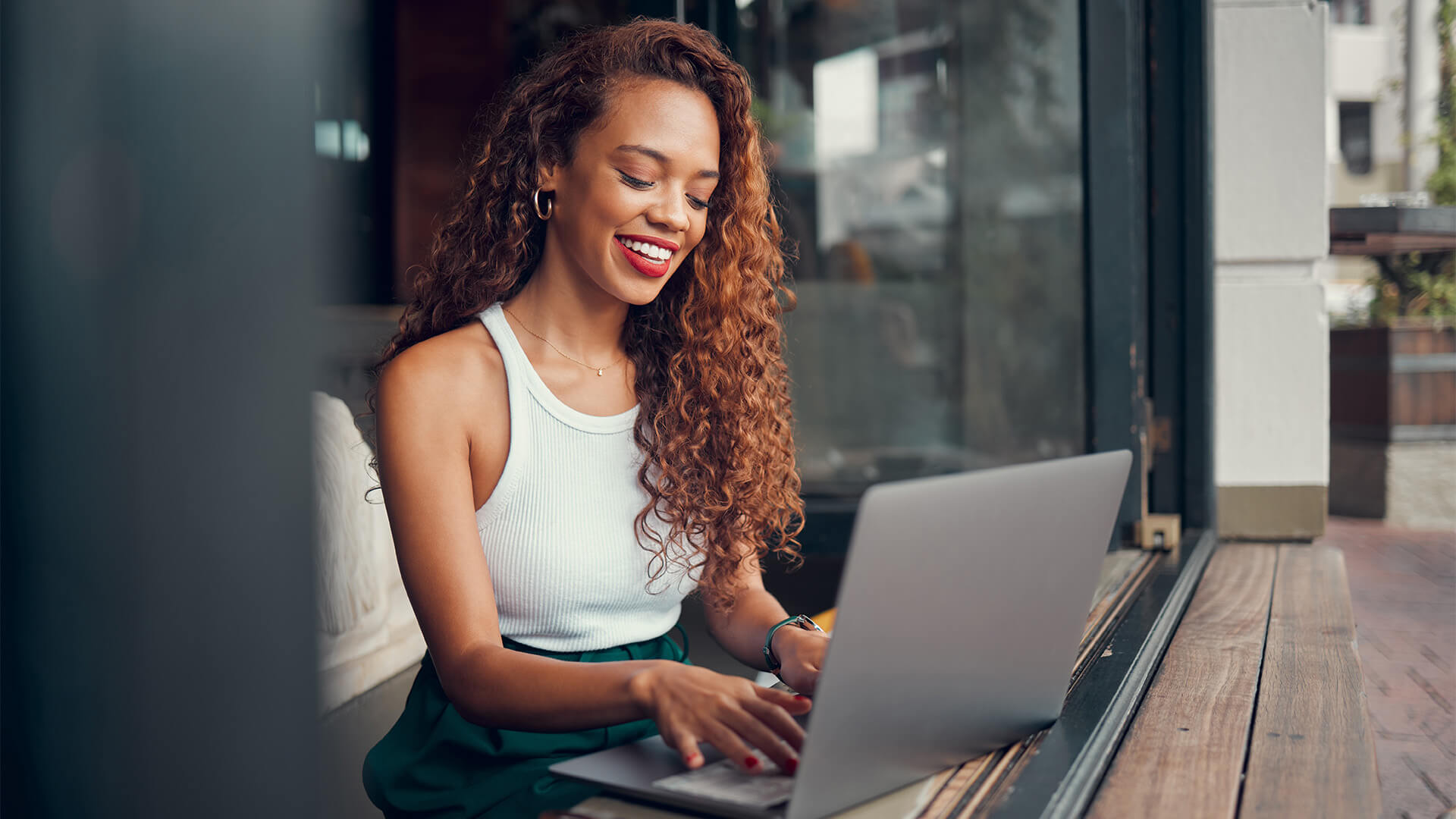 A woman in a white tank top and curly hair types on a laptop