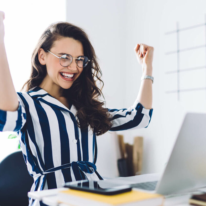 A woman in a blue and white blue striped blouse celebrates at a desk in front of herlaptop