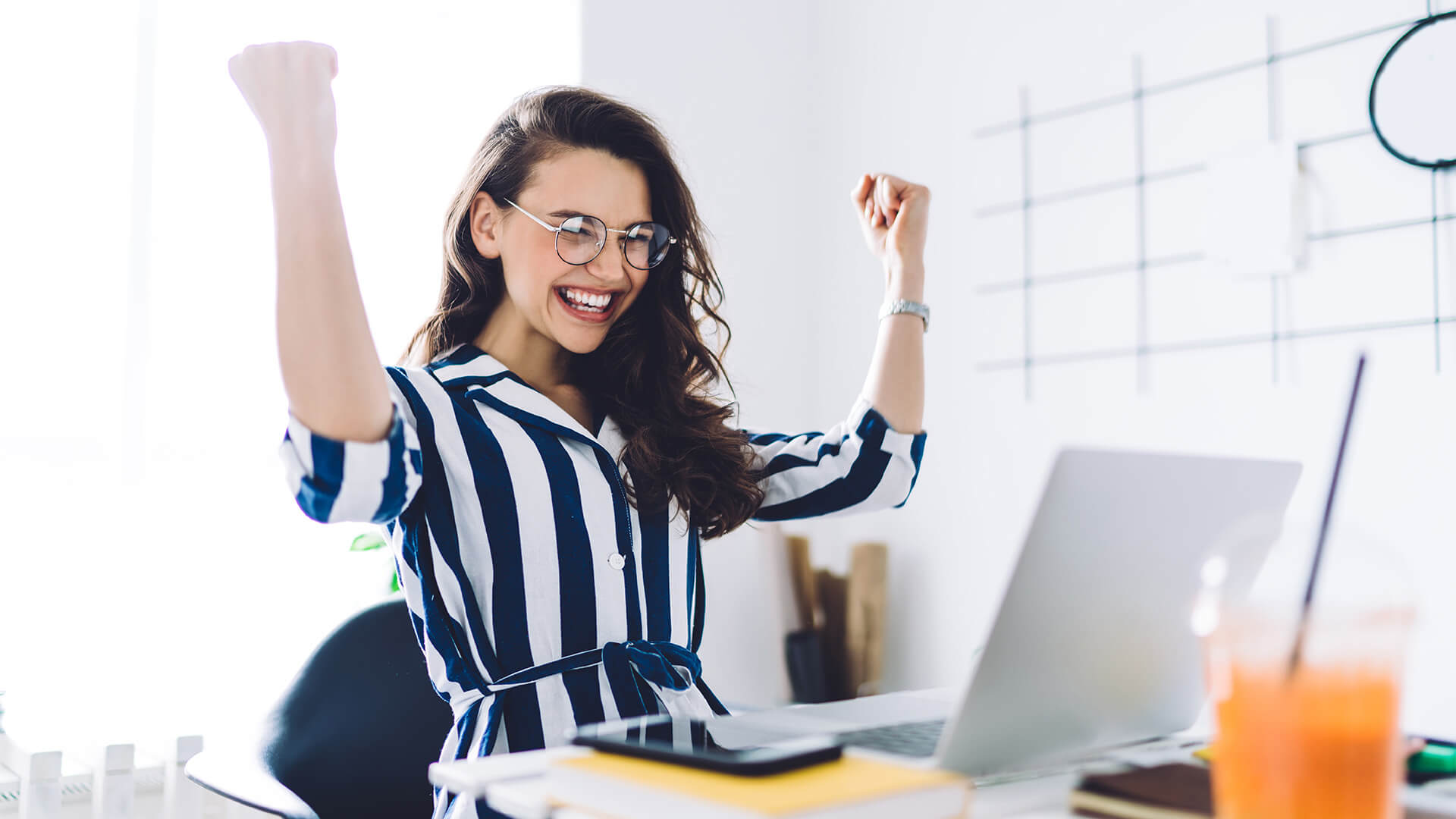 A woman in a blue and white blue striped blouse celebrates at a desk in front of herlaptop