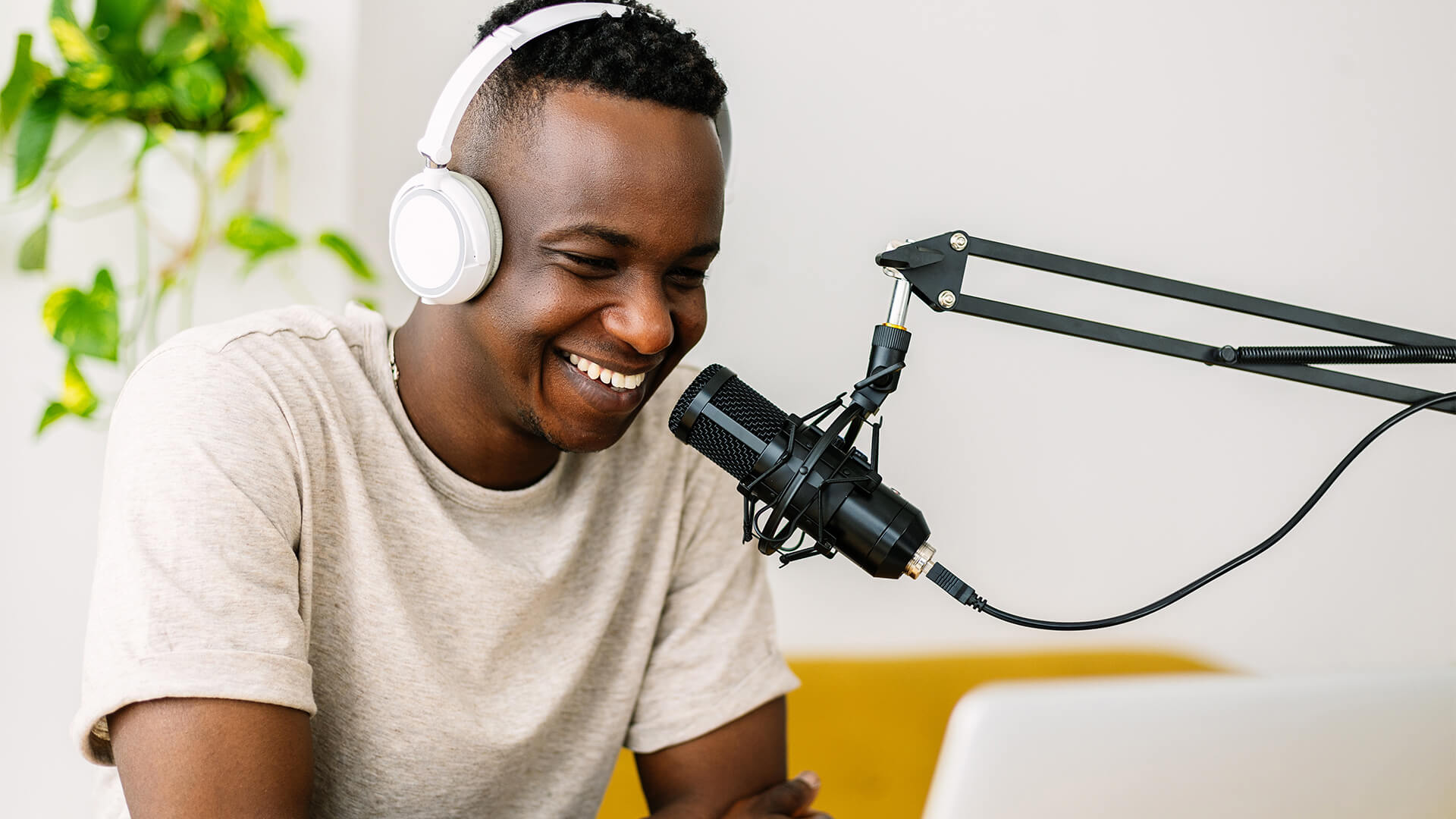 A man wearing white headphones smiles as he sits in front of a podcast microphone