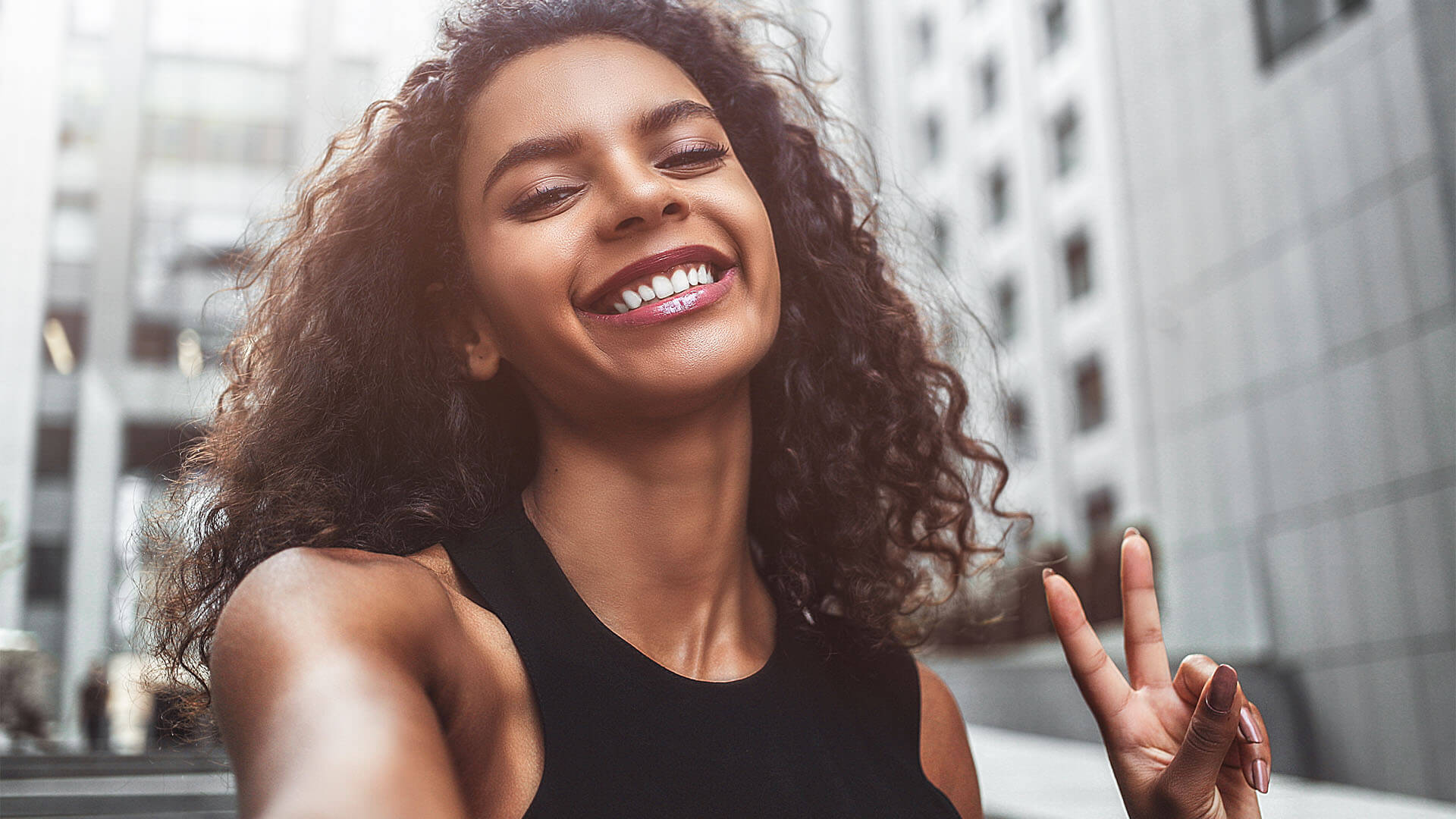 A woman with black curly hair holds up a peace sign; wearing a black tank and smiling