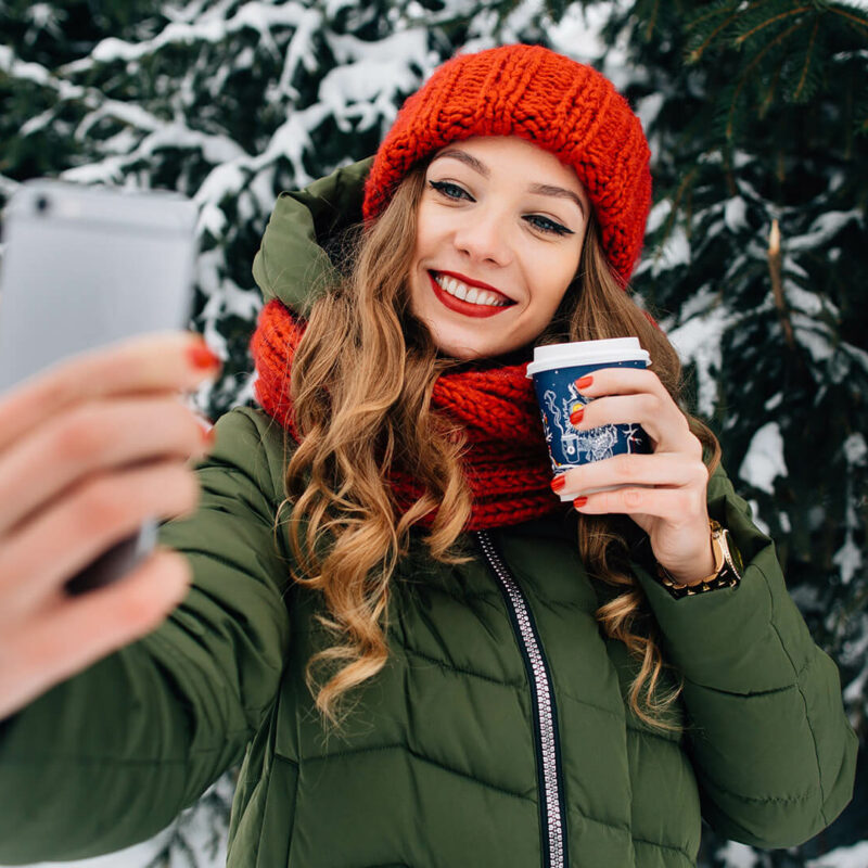 A woman with long brown curled hair and a red beanie and scarf holds a coffee cup while taking a selfie