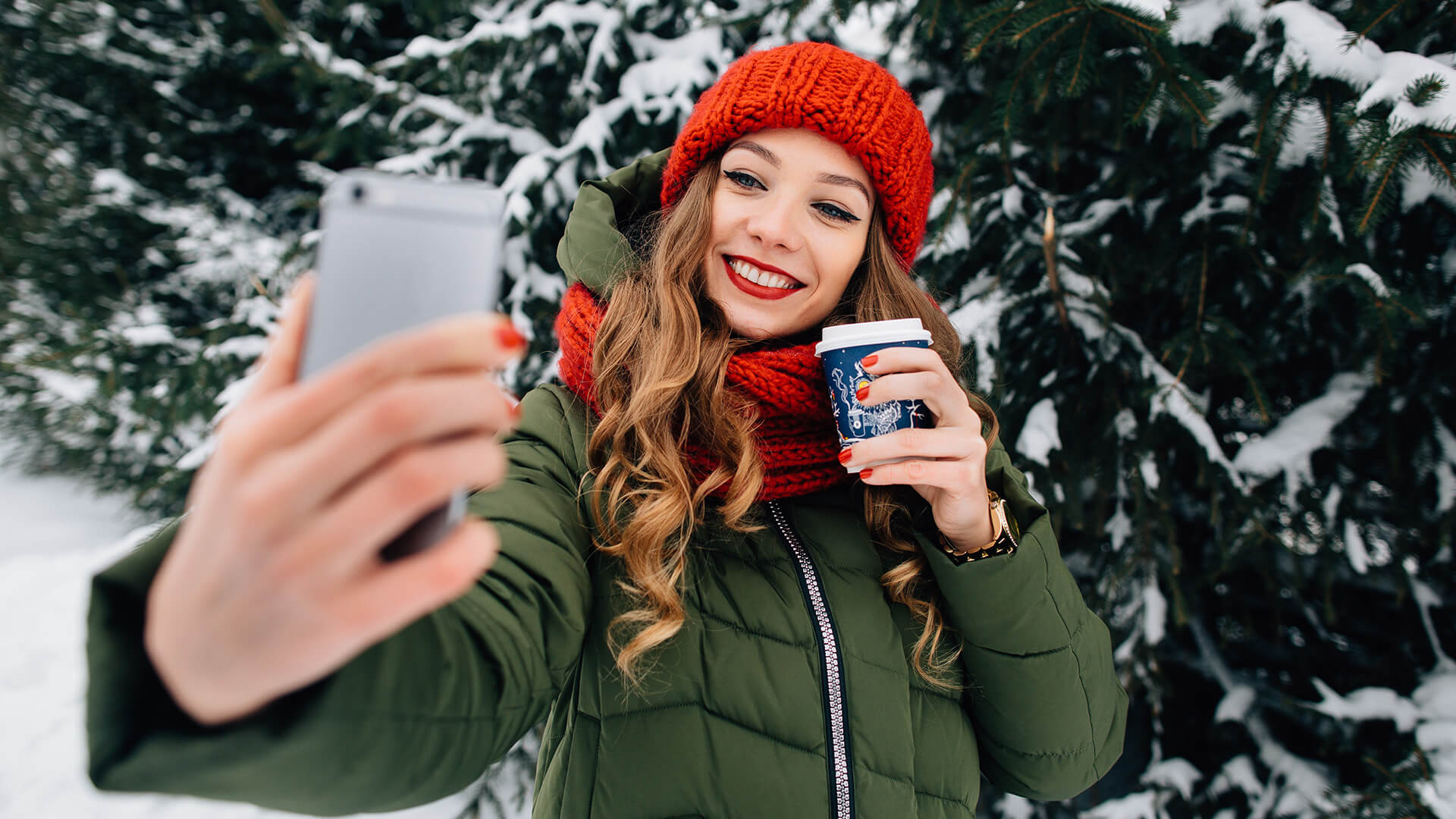 A woman with long brown curled hair and a red beanie and scarf holds a coffee cup while taking a selfie