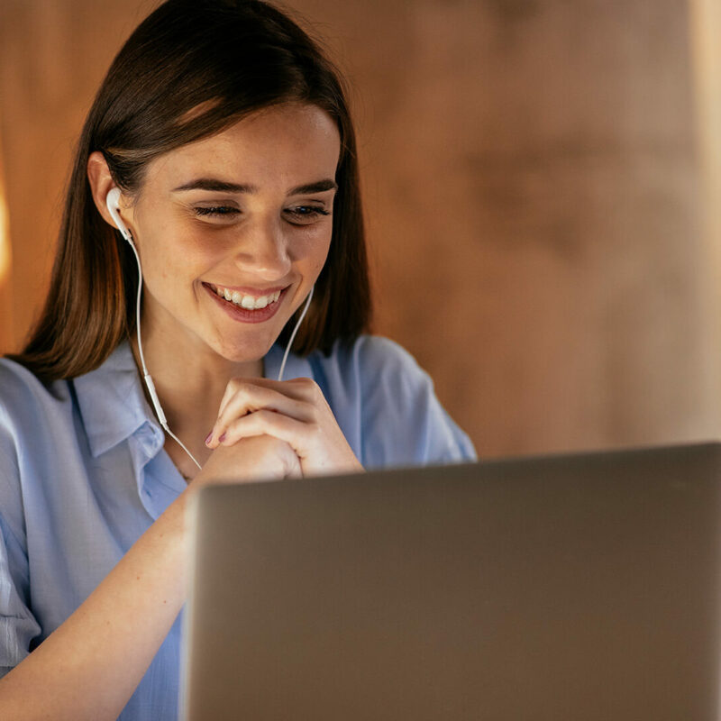 A woman with long brown hair smiles as she looks at her laptop screen; she has earphones in