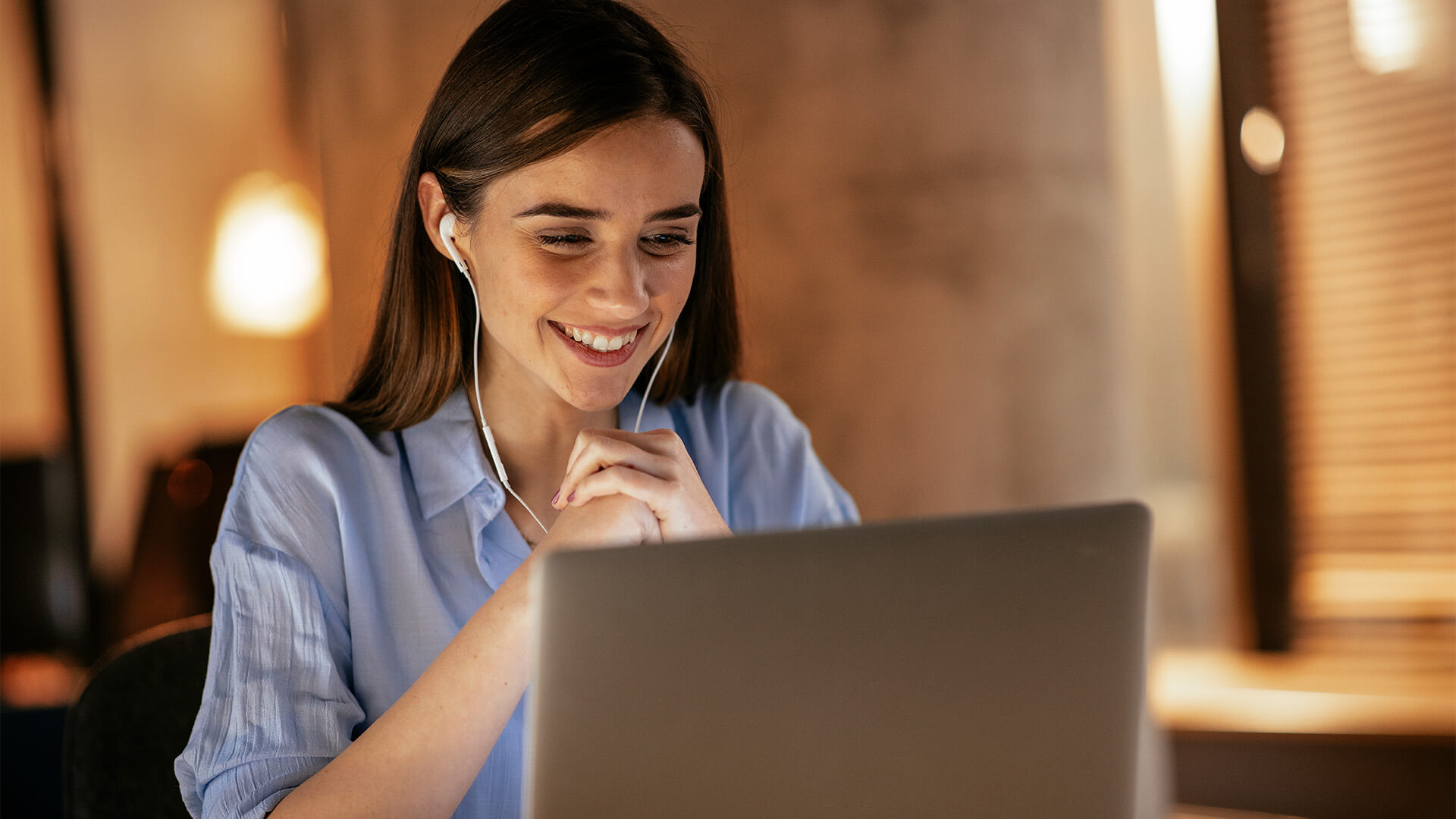 A woman with long brown hair smiles as she looks at her laptop screen; she has earphones in