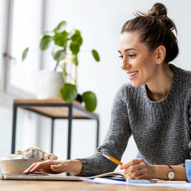 A woman in a dark gray sweater and hair in a bun types on a laptop while smiling