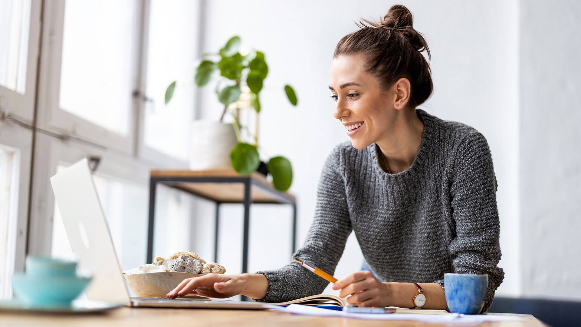 A woman in a dark gray sweater and hair in a bun types on a laptop while smiling