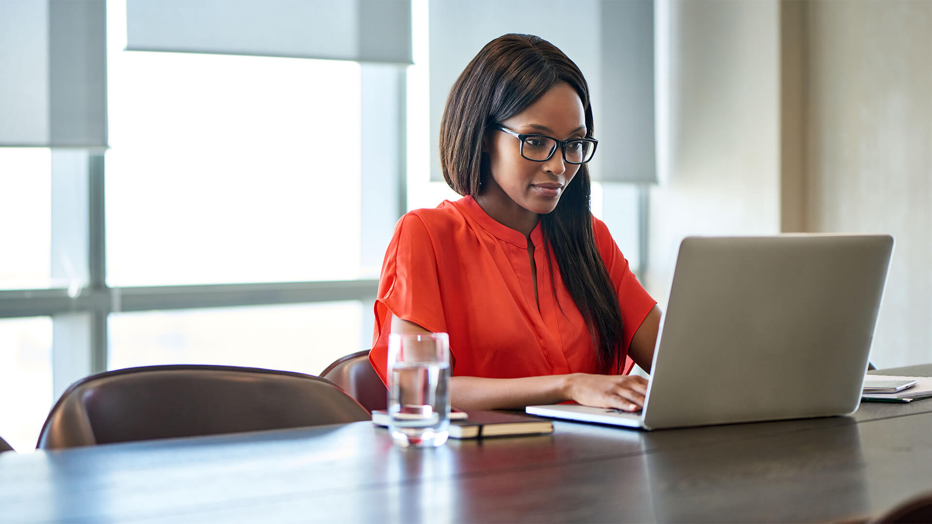 A Black woman wearing glasses and a red top types at her laptop in a conference room